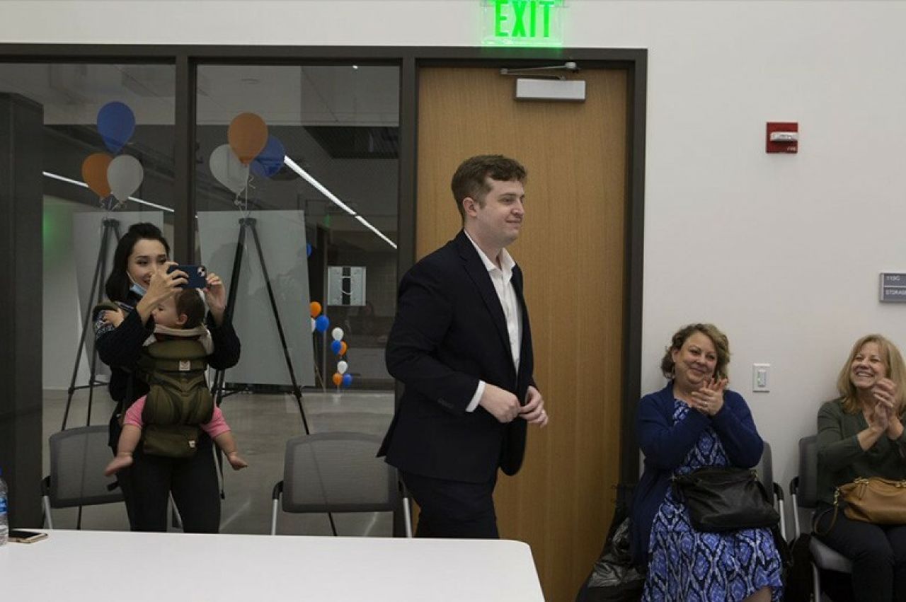 Young man with brown hair wearing a black suit walking to receive an ward