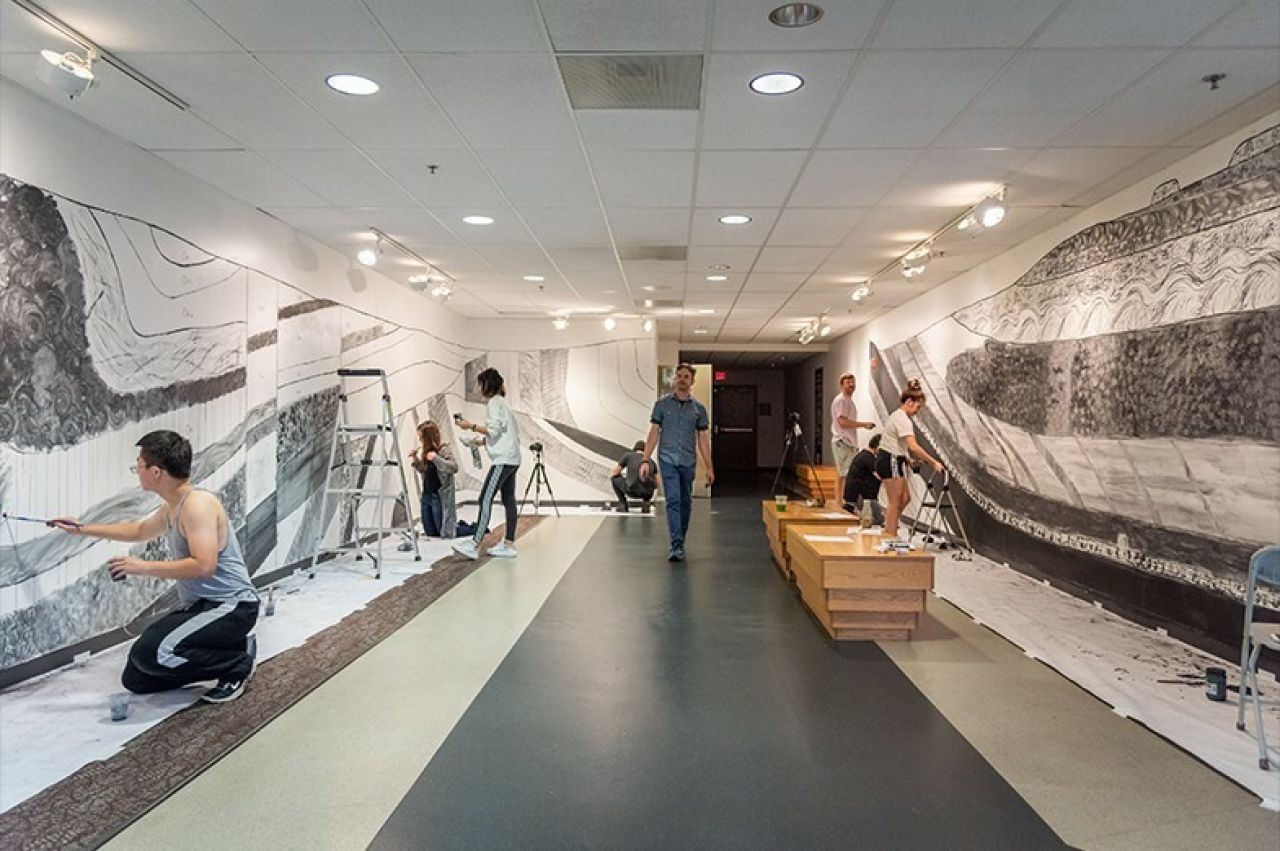 Alec Spangler walks towards the camera from the back of the gallery with students working on his wall drawing on either side of him.
