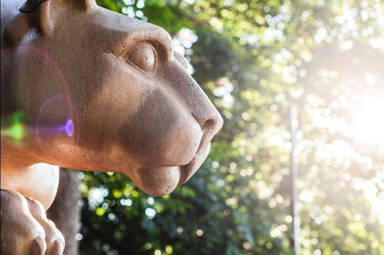 Profile of a light brown colored marble lion statue with a green tree background