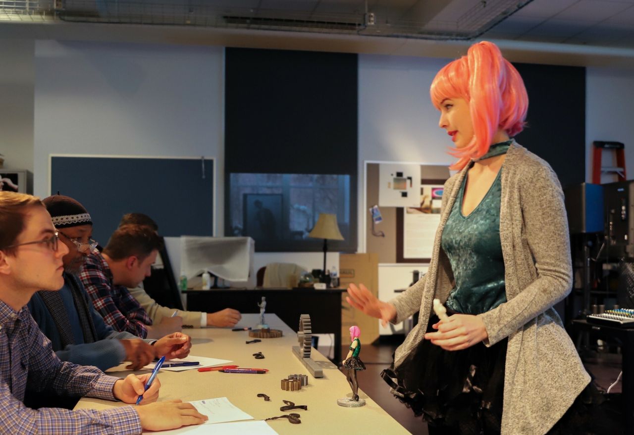 A student presents her 3d model figurine concept to a panel of judges during the Arts Business Idea Competition. Mimicking the look of her 3d model, she wears a pink ponytail wig and vibrant outfit.