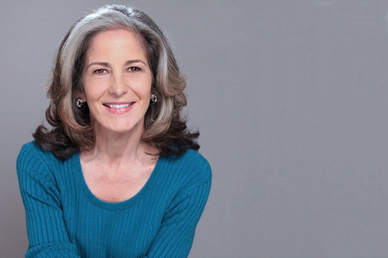 Photo of a woman with greying hair wearing a blue shirt and smiling in front of a grey background
