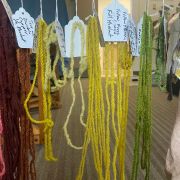 A series of colorful yarn hanks hanging from strings, each bit of yarn has a small handwritten tag with information about the dye used to color the yarn.