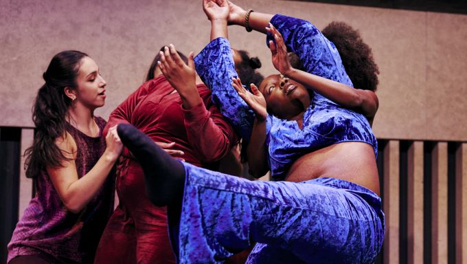 A small group of women of color cluster together in a pose supportive of one dancer.