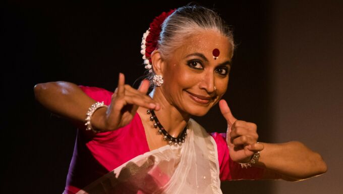 A woman in classical Indian dance dress gestures with her hands and fingers and smiles.