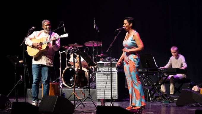 A man of color stands and plays guitar while a woman stands at a microphone and sings.