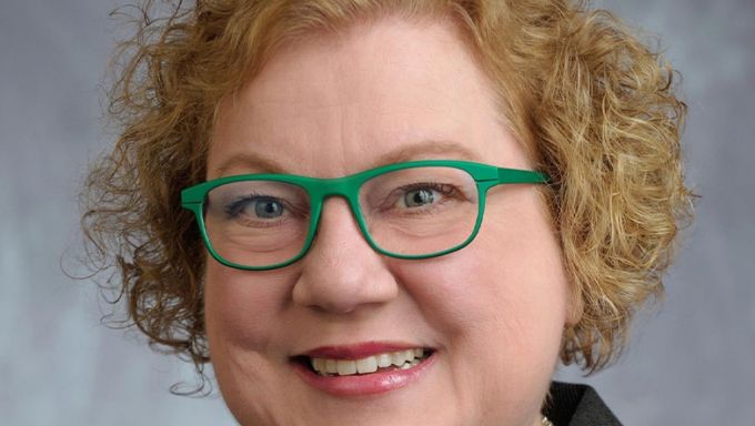 Head shot of a white woman with blonde curly hair and green-framed glasses smiling.