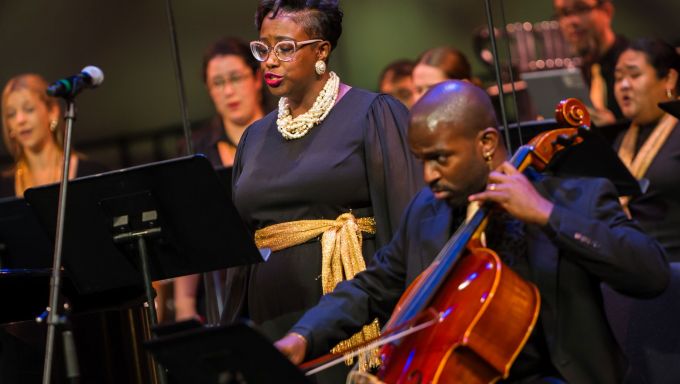 A diverse group of singers stand around a seated Black man playing cello.