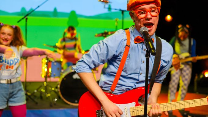 A boy wearing glasses plays a guitar in front of a diverse group of young people dancing behind him.