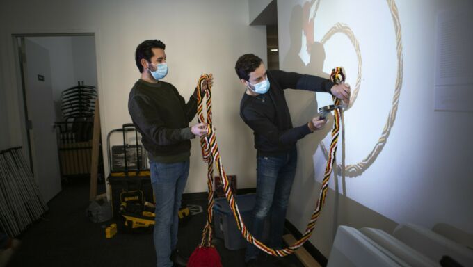 Two men attaching a colorful rope-like structure to a wall