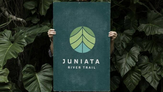 A woman holding a sign for the Juniata River Trail in front of her amid a background of plants.