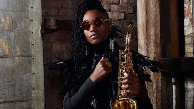 A Black woman wearing sunglasses stands against a brick wall and holds a shiny saxophone.