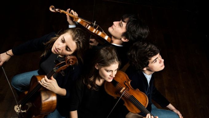 Four White young people stand together and hold their classical string instruments.