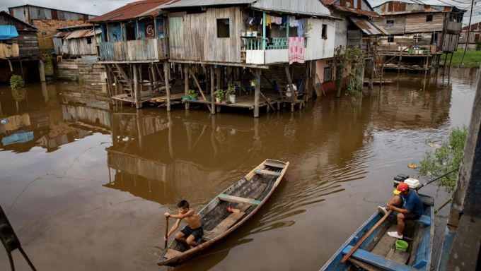 A young shirtless man rowing a boat through a floodplain community among houses on stilts in Iquitos, Peru.