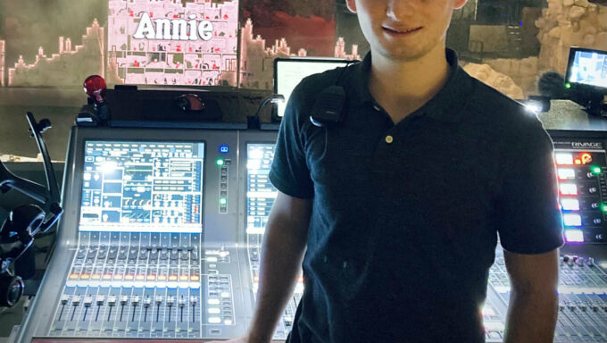 A young man stands in front of a sound production board with its knobs and buttons.