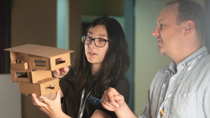 A summer camper at left holds an architectural model with an instructor at right.