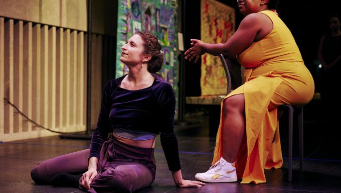 A white woman sits on a stage while a Black women sits in a chair next to her.