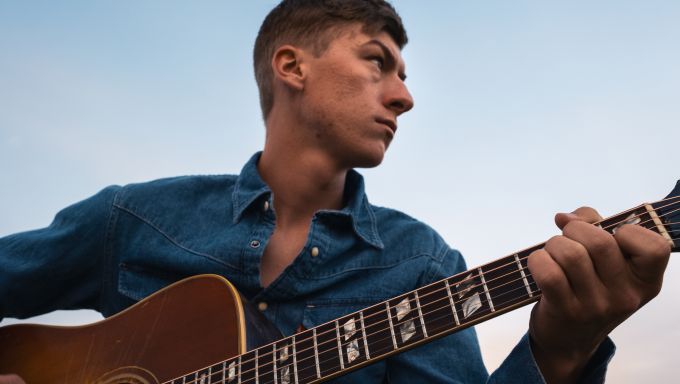 A young white man holds an acoustic guitar as he looks off into the distance.