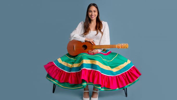 A smiling woman in a full skirt sits on a bench and holds a small guitar.