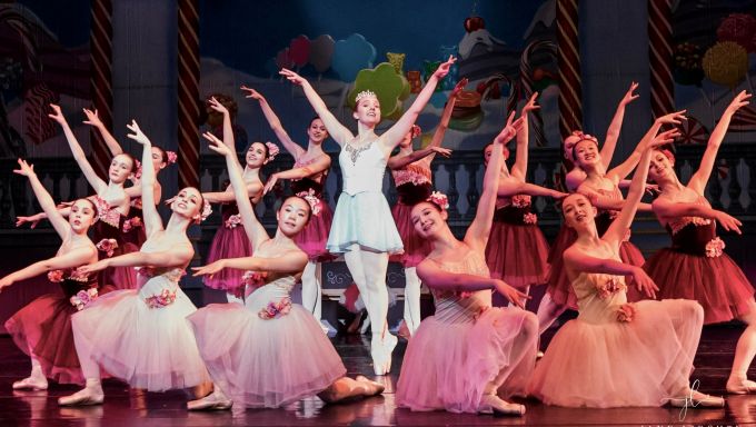 A group of young ballet dancers wear matching tutus and crowd around a dancer in the center in the pointe position.