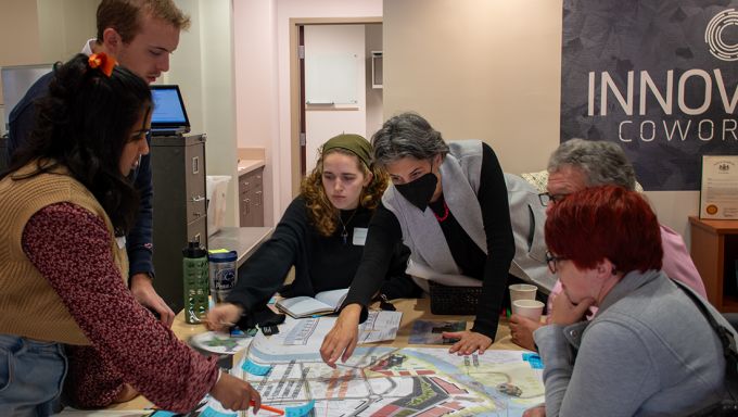 A woman leans across a table to point at a map as students and community members at the table look down at the mapl