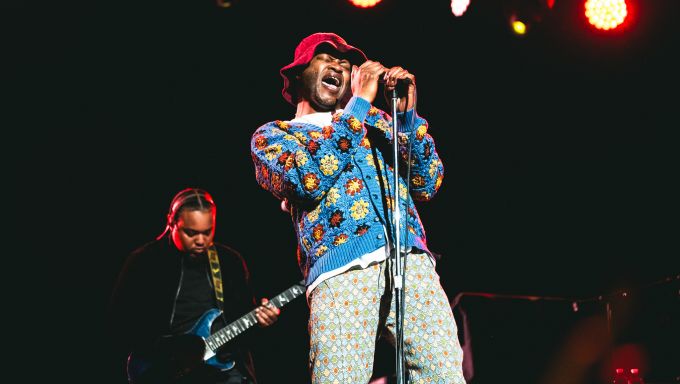 A black man wearing a crocheted suit sings passionately into a microphone on a darkened stage.