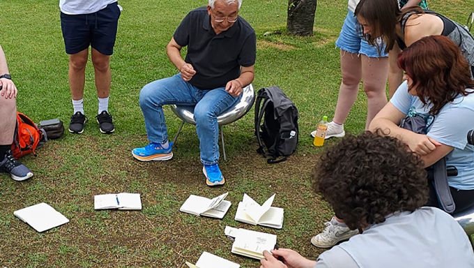 A man sitting in a chair outsides, surrounded by students, with sketchbooks open on the ground in front of them.