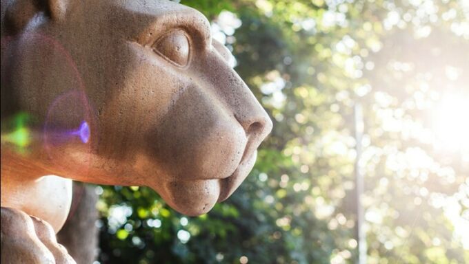 Profile of a light brown colored marble lion statue with a green tree background