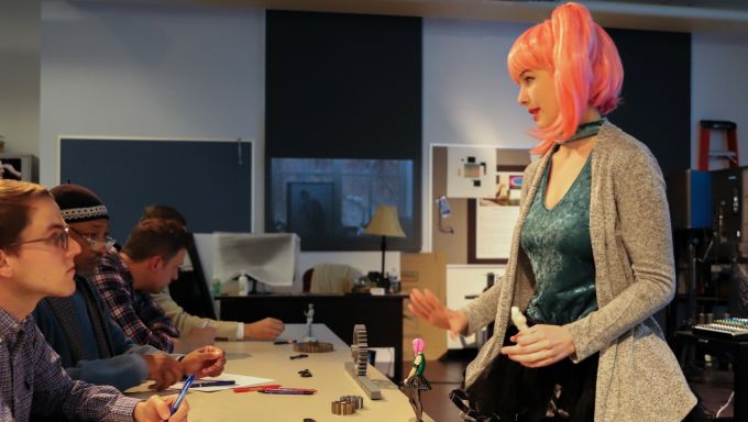 A student presents her 3d model figurine concept to a panel of judges during the Arts Business Idea Competition. Mimicking the look of her 3d model, she wears a pink ponytail wig and vibrant outfit.