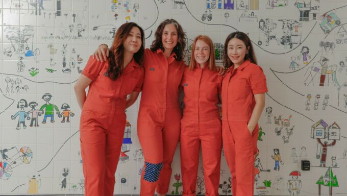Four women in orange jumpsuits in front of a mural