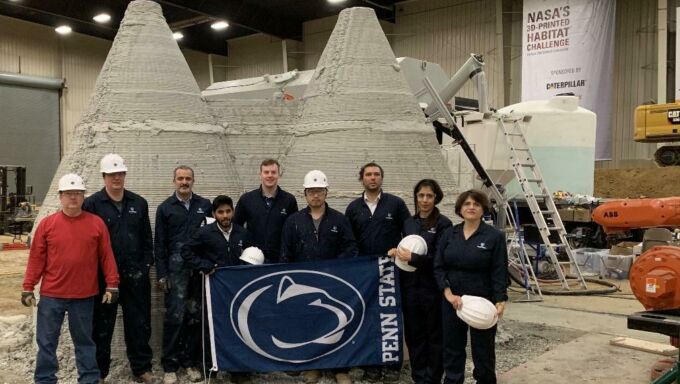 Penn State NASA Mars Challenge team standing together in front of full-size 3d-printed concrete dwellings holding Penn State flag