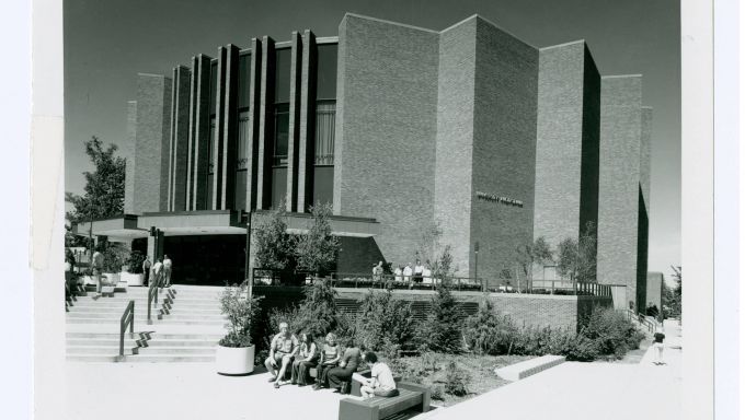 An archival photo shows the large, brick Eisenhower Auditorium and its stepped patio, landscaping and tall vertical windows.