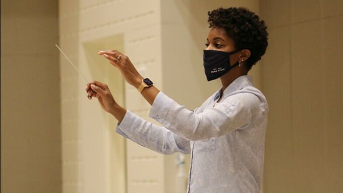 African American woman with short, dark hair wearing a blue face mask and a white long-sleeved shirt conducts a choir
