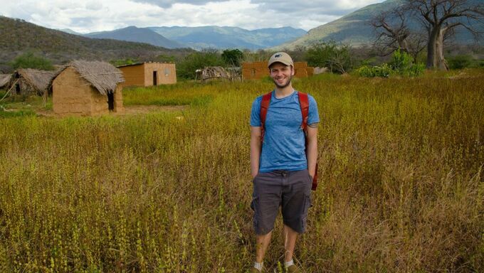 Man in blue shirt and grey shorts standing in front of grass-thatched huts with the mountains of Tanzania towering in the background.