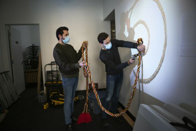 Two men attaching a colorful rope-like structure to a wall