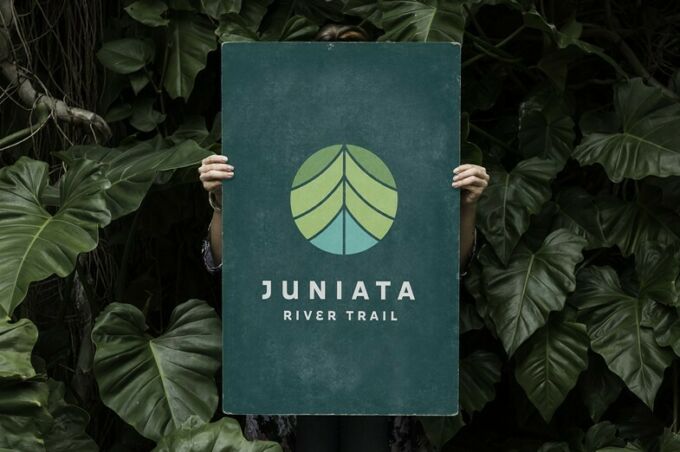 A woman holding a sign for the Juniata River Trail in front of her amid a background of plants.