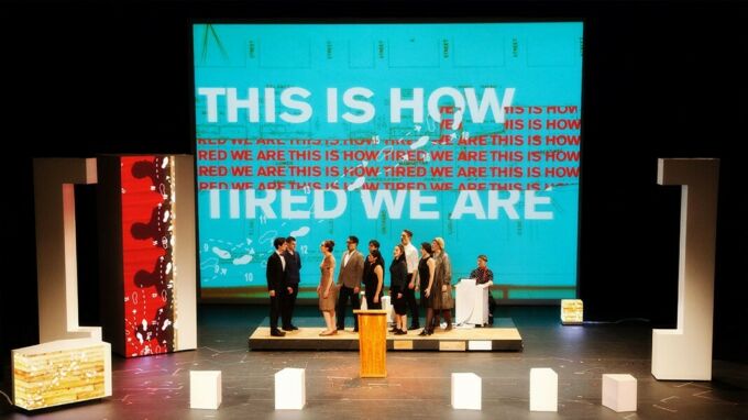 A group of people cluster together in front of a screen with the words "This is how tired we are."