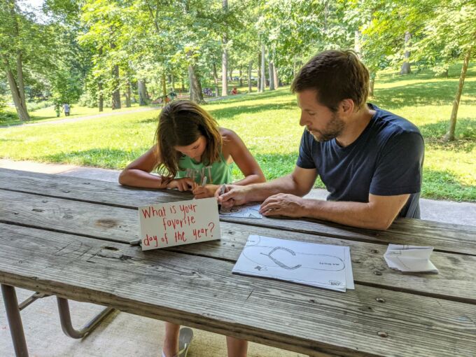 A man helps a young girl with a drawing at a table outdoors. A table tent in front of them reads, "What is your favorite day of the year?"