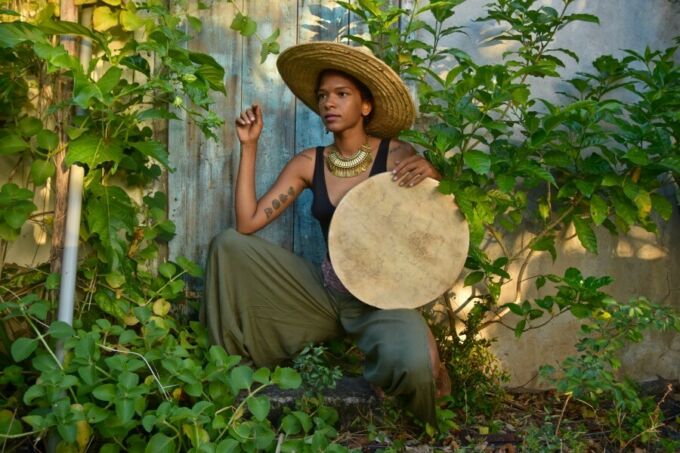 A woman wears a hat and holds a drum as she sits amid trees.