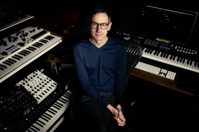 A man wears round black-rimmed glasses and sits surrounded by high-tech keyboards and synthesizers.