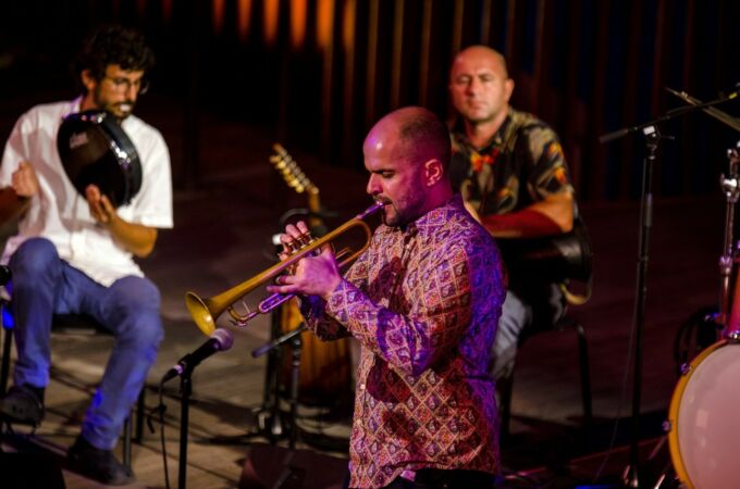 A man plays a trumpet while two percussion musicians in the background accompany.