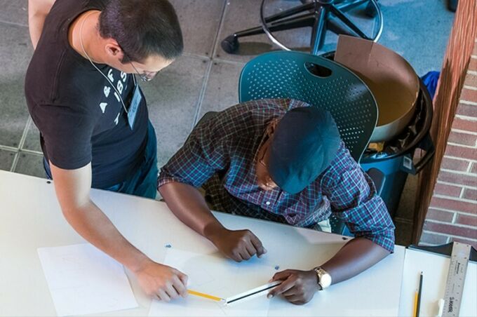 A faculty member instructs an architecture student as he sits at his drawing table.