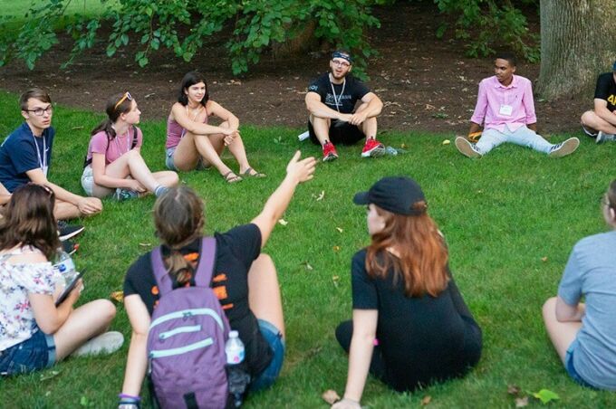 Summer camp participants sitting in a circle on a patch of grass talking.