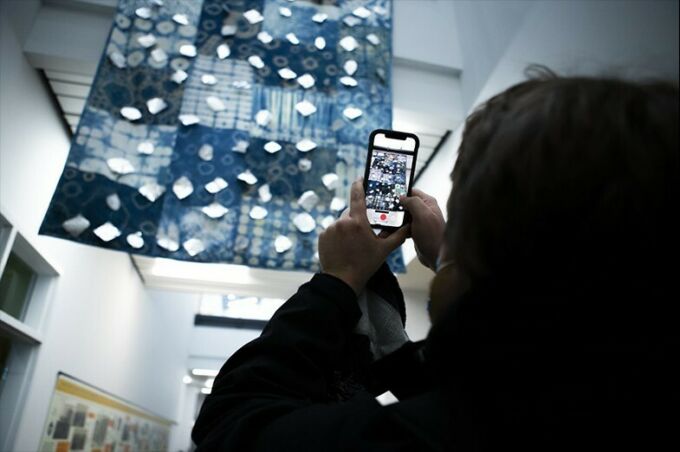 A person takes a photo of the indigo-colored quilt hanging above them with their smartphone.