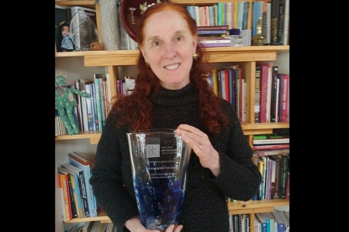 Woman with mid-length red hair holding a glass vase award with a book shelf background