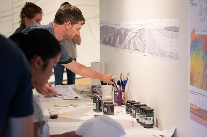 Alec spangler and students at left grab brushes and graphite from a table in front of them with a geological map on the wall above it.
