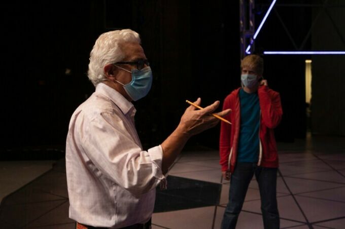 A white man with light grey hair, black rimmed glasses and a face mask in a white shirt directs actors