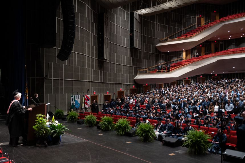 A master of ceremonies standing at a podium on a stage addresses a seated audience.