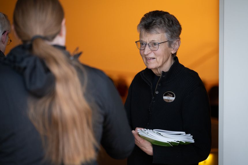 An older woman wearing a crisp uniform stands and offers event programs to passersby.