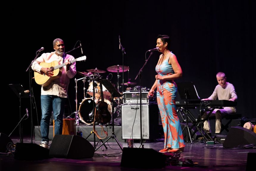 A man of color stands and plays guitar while a woman stands at a microphone and sings.