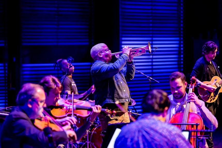 A man of color standing in the center of seated string musicians plays a trumpet.